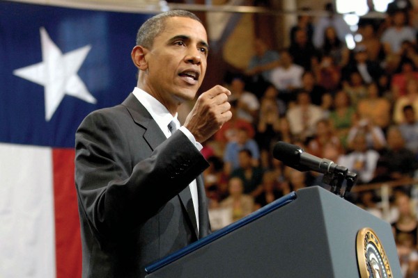 A man speaking at a podium with the Texas flag and a crowd of people in the background