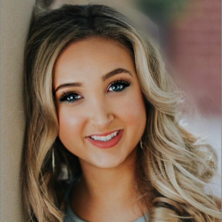 A younger woman with longer blond hair looking at the camera and leaning her head against a wall