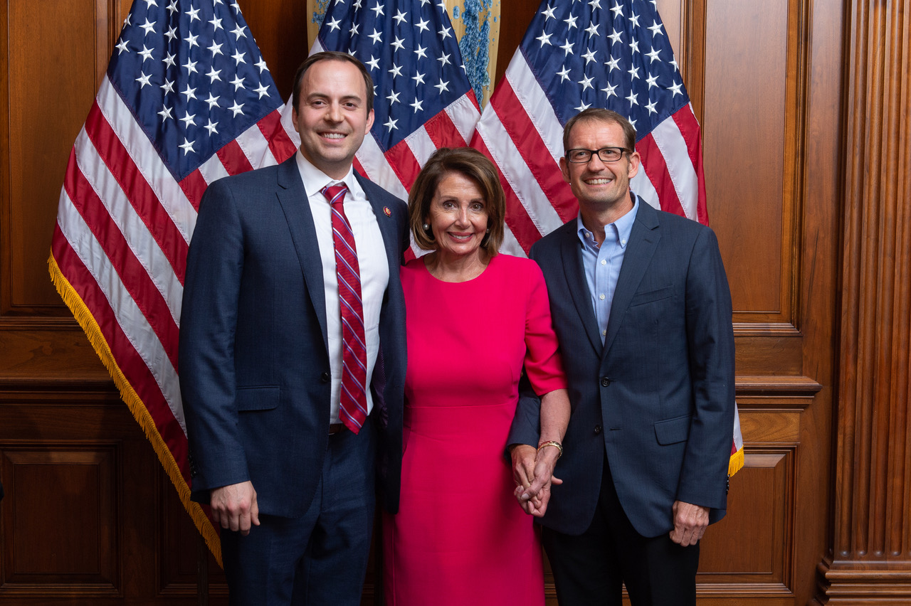 Rep. Lance Gooden, Speaker Nancy Pelosi, and Professor Sean Theriault during Gooden's swearing-in ceremony with three American flags and wooden panel walls as background.