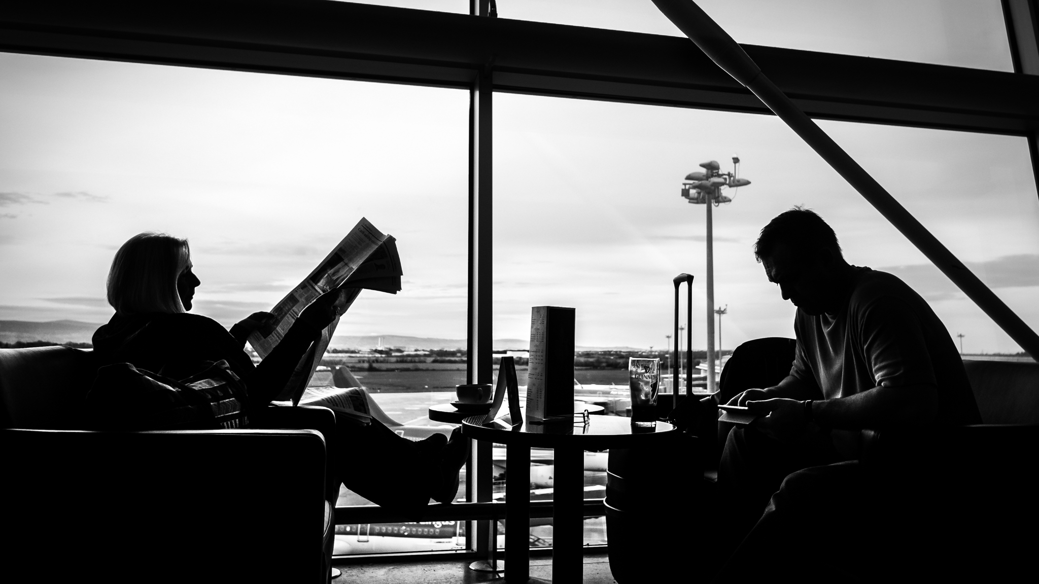 At a large window, a woman reads a newspaper and a man reads on his phone. They are on chairs across a table from each other, and the photo shows their profiles.
